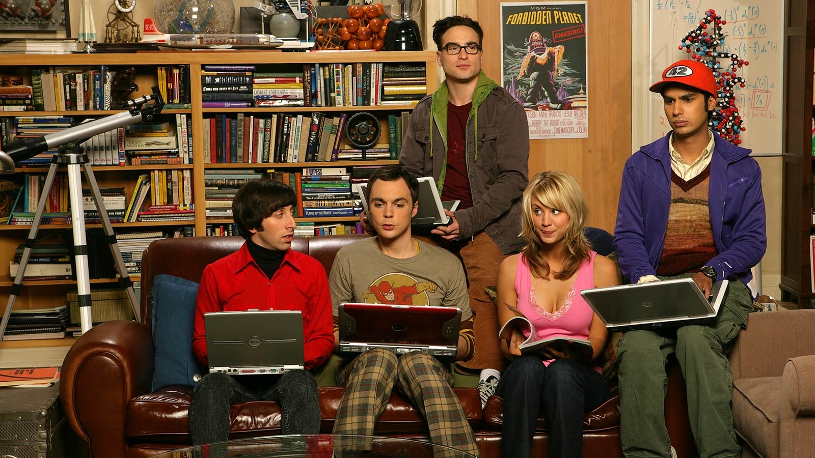 Quatro homens com laptops e uma mulher com uma revista sentados em um sofá. Os homens vestem roupas de super-heróis e têm uma decoração com muitos livros, pôsteres, equações matemáticas e ferramentas científicas.